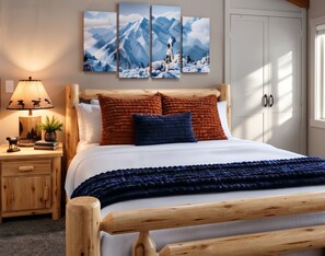 A bedroom with a wooden bed frame, white bedding, a blue blanket, and a mountain-themed wall art above the bed.
