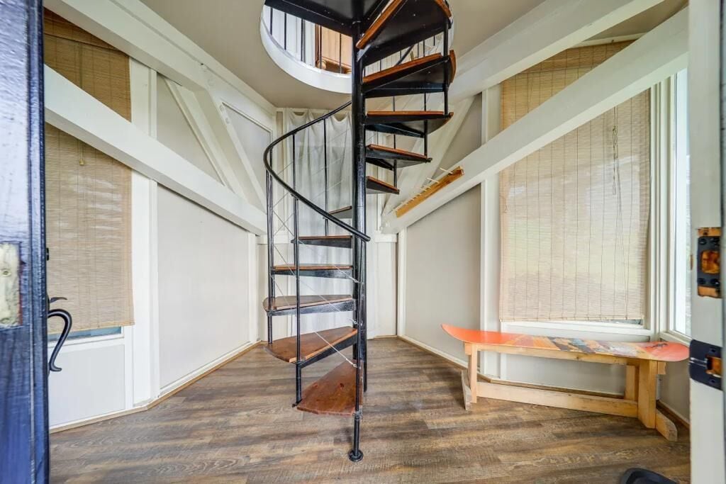 Una escalera de caracol en una habitación con suelo de madera, ventanas amplias con persianas y un banco junto a la ventana.