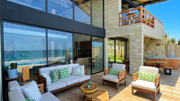 A modern outdoor living area with a wooden deck, large windows overlooking the ocean, and a second-story balcony with a wooden railing. - DIEM Vichayito Beachfront Eco-Luxury (Los Organos)