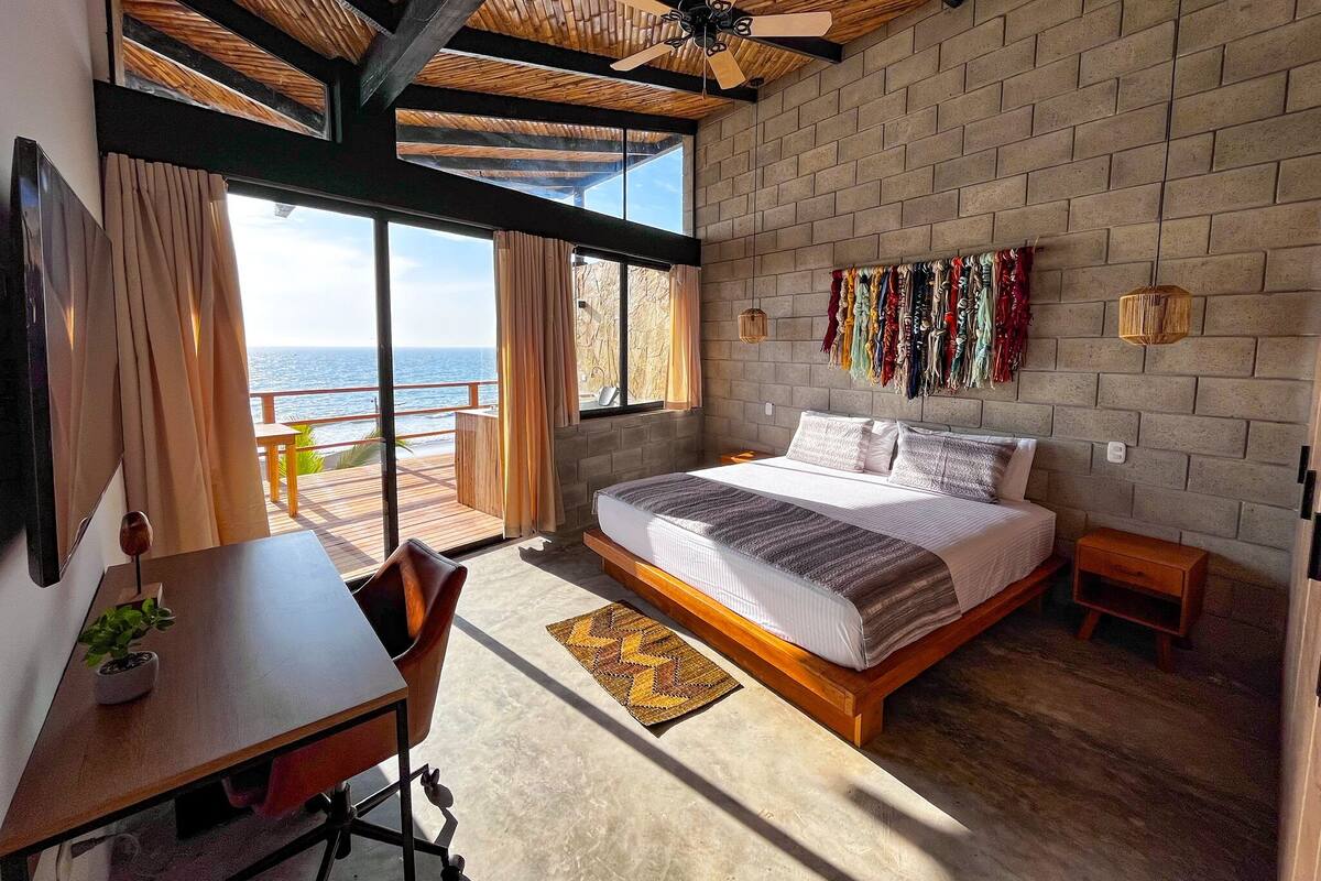 Une chambre avec un grand lit, un bureau et une chaise. De grandes fenêtres offrent une vue sur l’océan.