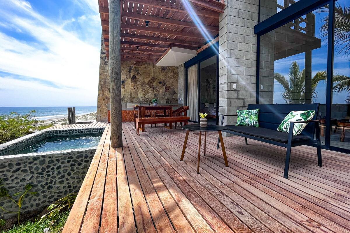 Une terrasse en bois avec une piscine bordée de pierres, une table en bois et un canapé avec des coussins verts.