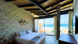 A bedroom with a large bed, a wooden headboard, a wall-mounted decorative item, a nightstand, and a view of the beach through a large window. - DIEM Vichayito Beachfront Eco-Luxury (Los Organos)