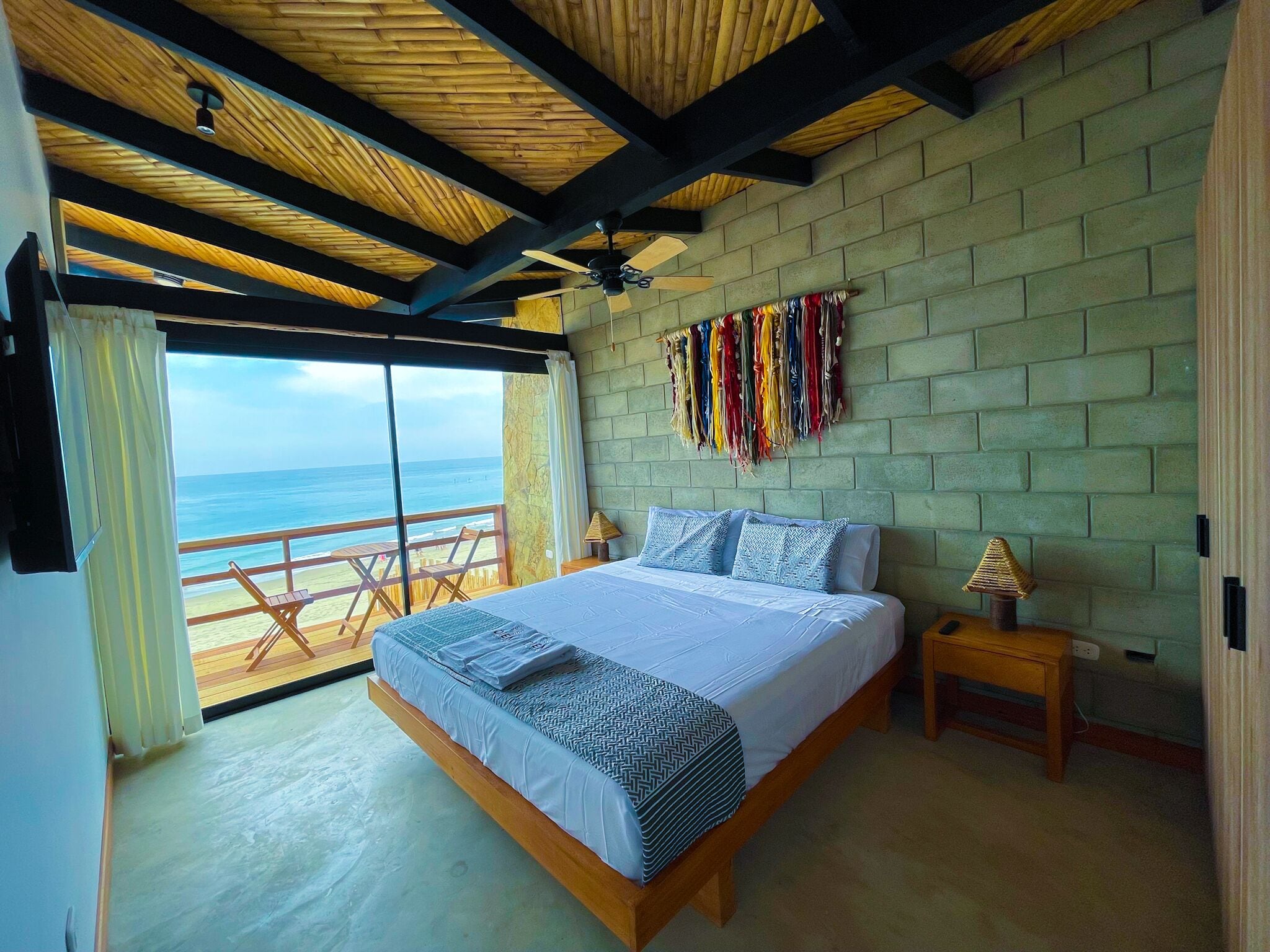 A bedroom with a large bed, a wooden ceiling, a view of the ocean, and a brick wall.
