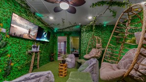 Une pièce avec un mur vert, un ventilateur de plafond, une télévision, des fauteuils en forme de poire, une chaise suspendue et une petite table avec une plante.