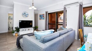 Een moderne woonkamer met een grijze bank, een flatscreen televisie en een eethoek met een tafel en stoelen.