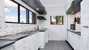 Een moderne keuken met witte kasten, een zwart aanrecht en een betegelde vloer. Er zijn ramen met uitzicht op palmbomen en een stadsbeeld.