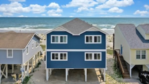 Een pand aan het strand met een twee verdiepingen tellend huis, voorzien van een balkon, een overdekte veranda en een trap naar een tweede niveau.