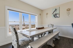 A dining area with a large window, a white table, and benches. There are wall decorations including a pelican painting and a flower.