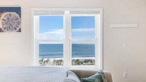 Een slaapkamer met een groot raam met uitzicht op de oceaan, een bed met een gestructureerde witte hoes en een lichtschakelaar aan de muur.