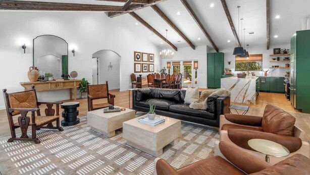 A spacious living area with a high ceiling, exposed beams, a black leather sofa, wooden chairs, and a kitchen in the background. - 6 Mins from Main St, Hot tub, Chef's Kitchen! (Fredericksburg)
