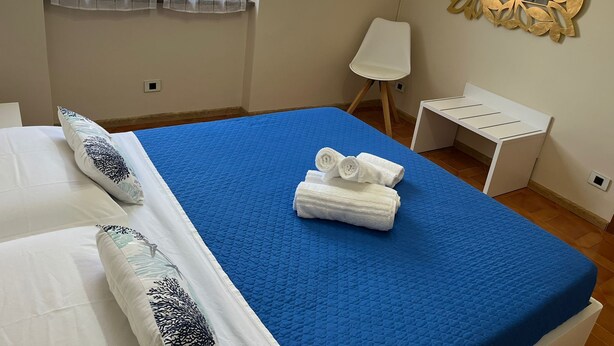 A hotel room with a bed, a blue bedspread, a white bedside table, a white chair, and a decorative mirror on the wall. - Valentina's Rent Apartment (Fertilia)