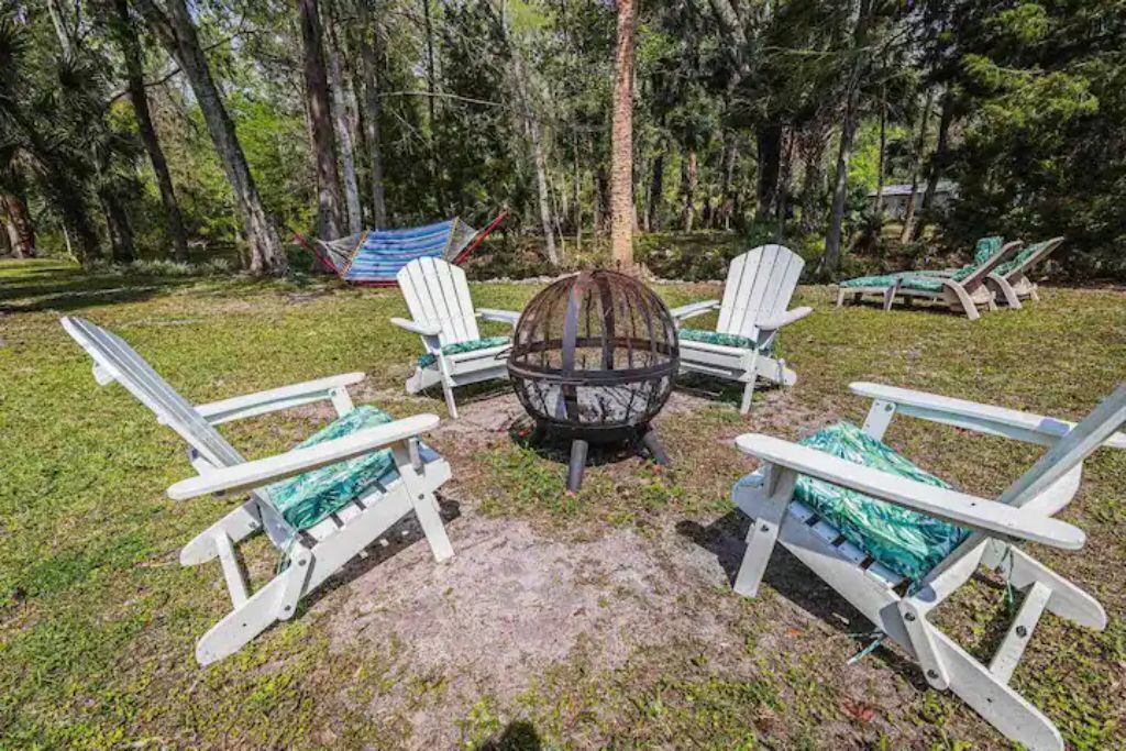 A circular seating arrangement with a central fire pit, surrounded by four white Adirondack chairs with green cushions, set in a grassy area with trees in the background.