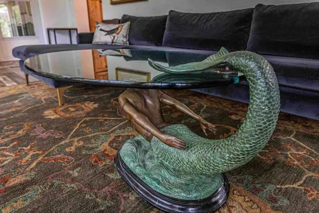 En grøn drageskulptur på et glastop med en sort fod, placeret på et mønstret tæppe i et rum med en mørkeblå sofa og et lille bord i baggrunden.