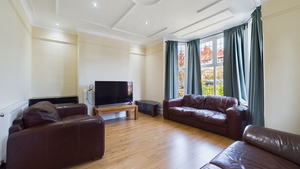 A spacious living room with a flat-screen TV, leather sofas, wooden flooring, and large windows with curtains.