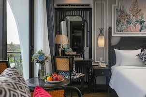 Habitación de hotel con una cama, un escritorio con lámpara, una silla, una mesa con fruta y un cuadro en la pared.