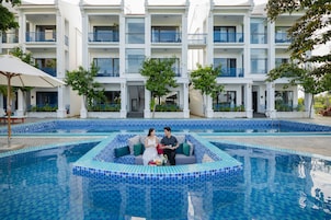 Un área de piscina con bordeado de azulejos, un edificio blanco con balcones azules, y una pareja sentada en un banco.