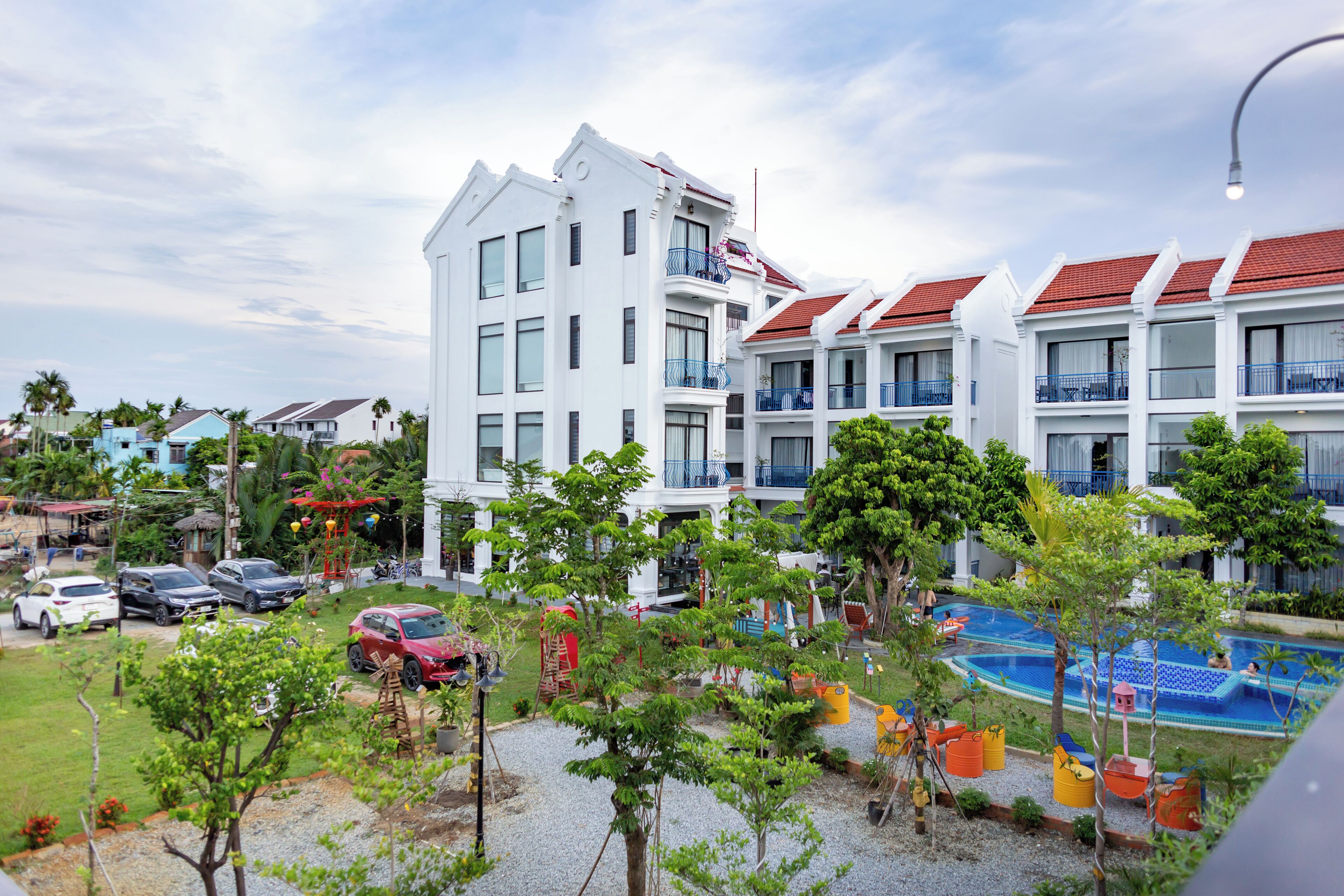 A hotel with multiple white buildings, a swimming pool, and a parking area with cars.