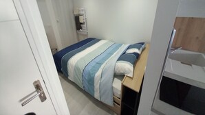 Een kleine, verzorgde slaapkamer met een bed, een houten nachtkastje, een ingebouwde heater en een deur.
