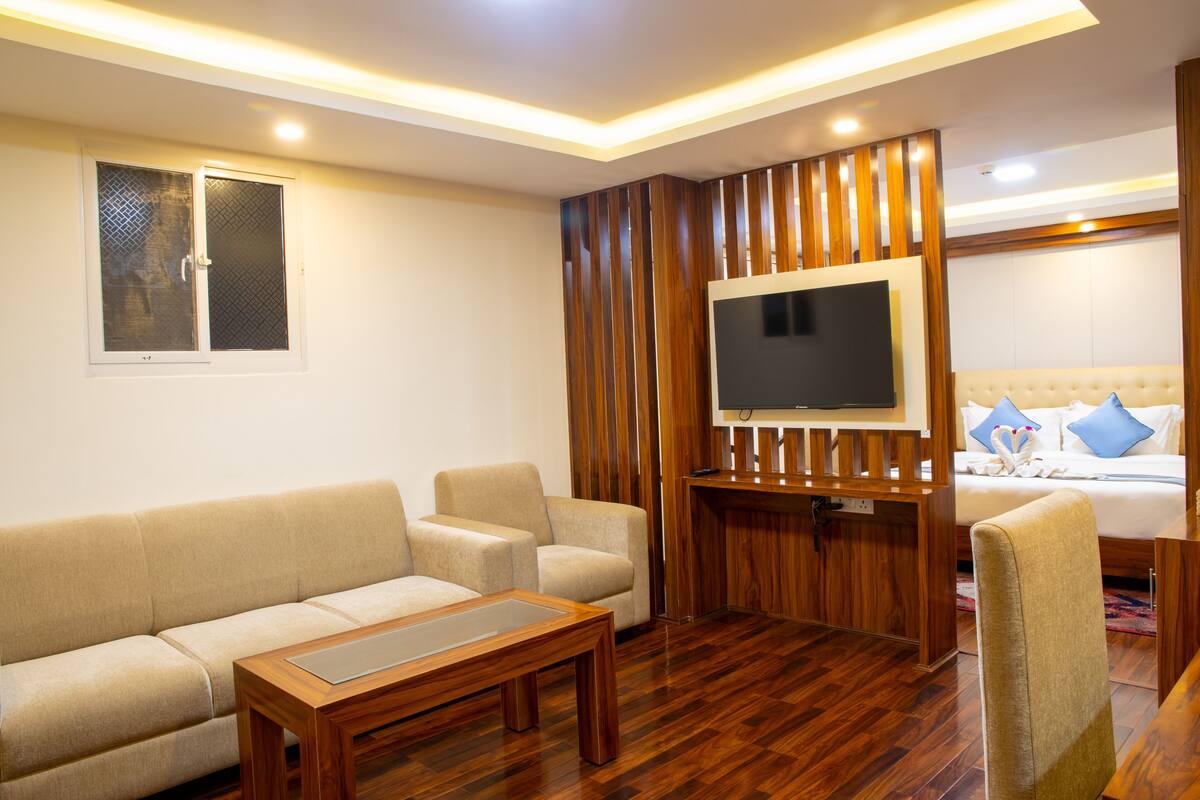 A modern living room with a sofa, a wooden coffee table, a TV mounted on a wooden panel, and a bed with blue pillows.