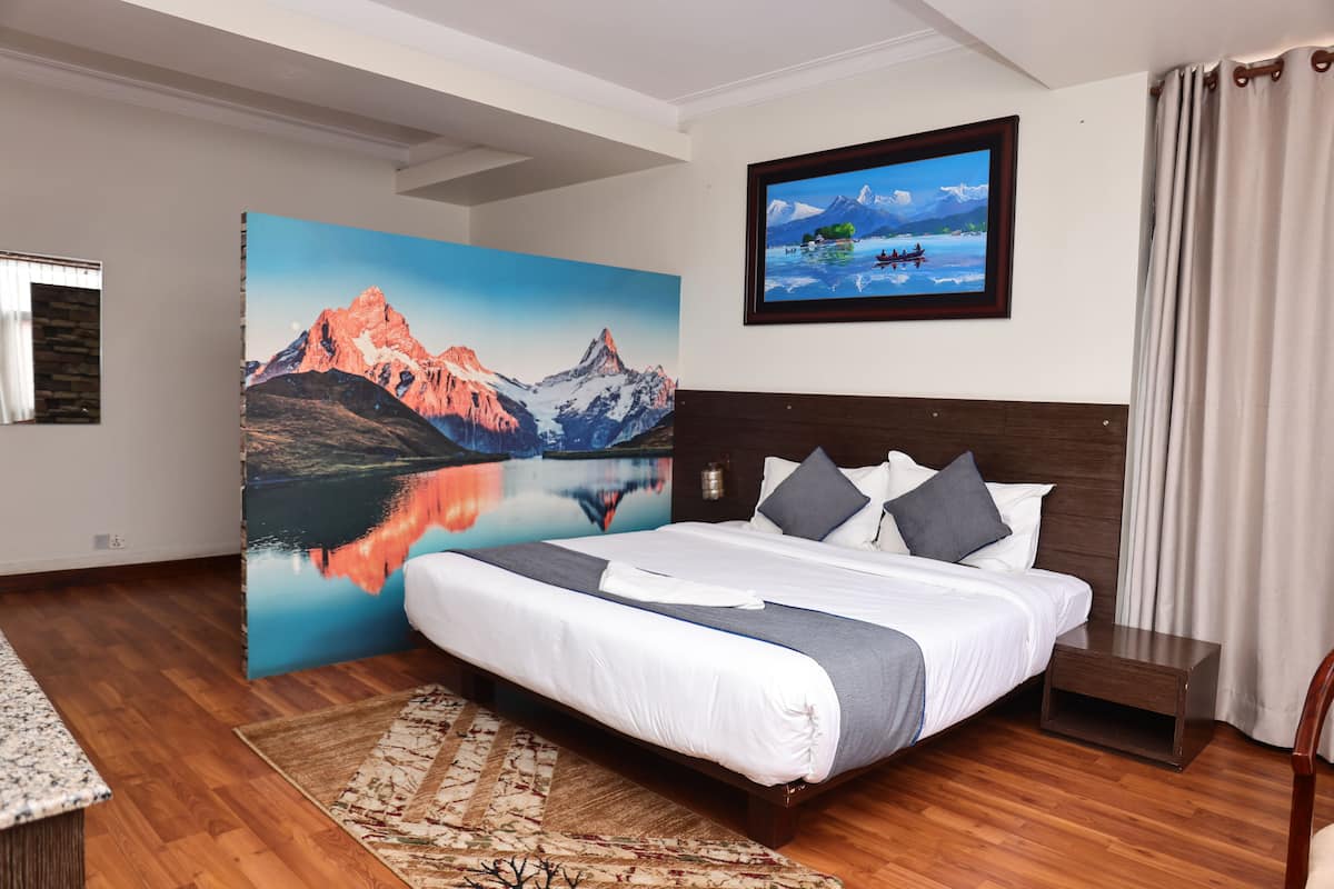 A hotel room with a large bed, a painting of a mountain landscape, a television on the wall, and a wooden floor.