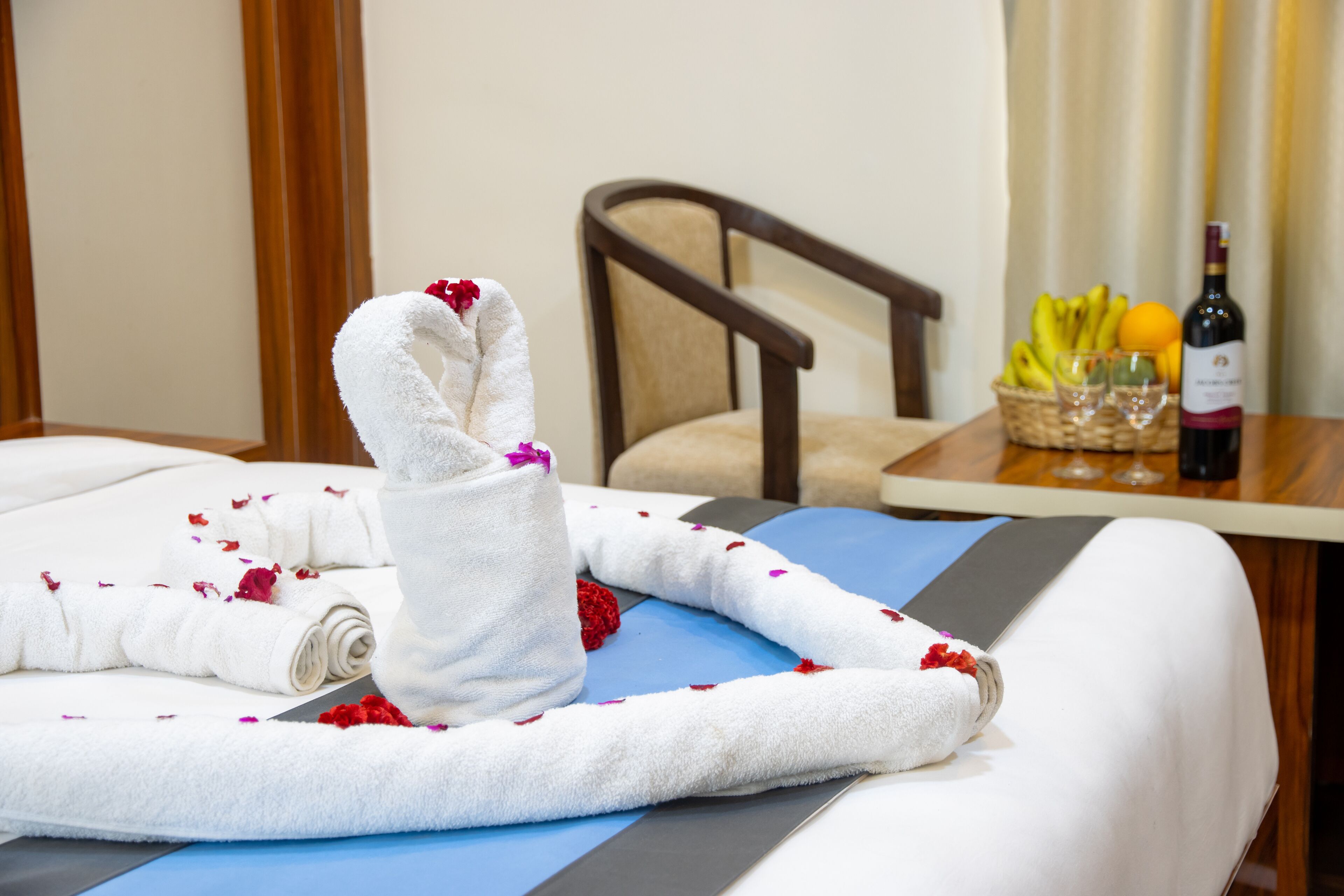 A neatly made bed with a towel arranged in a heart shape, a bottle of wine, and a basket of fruit on a table.