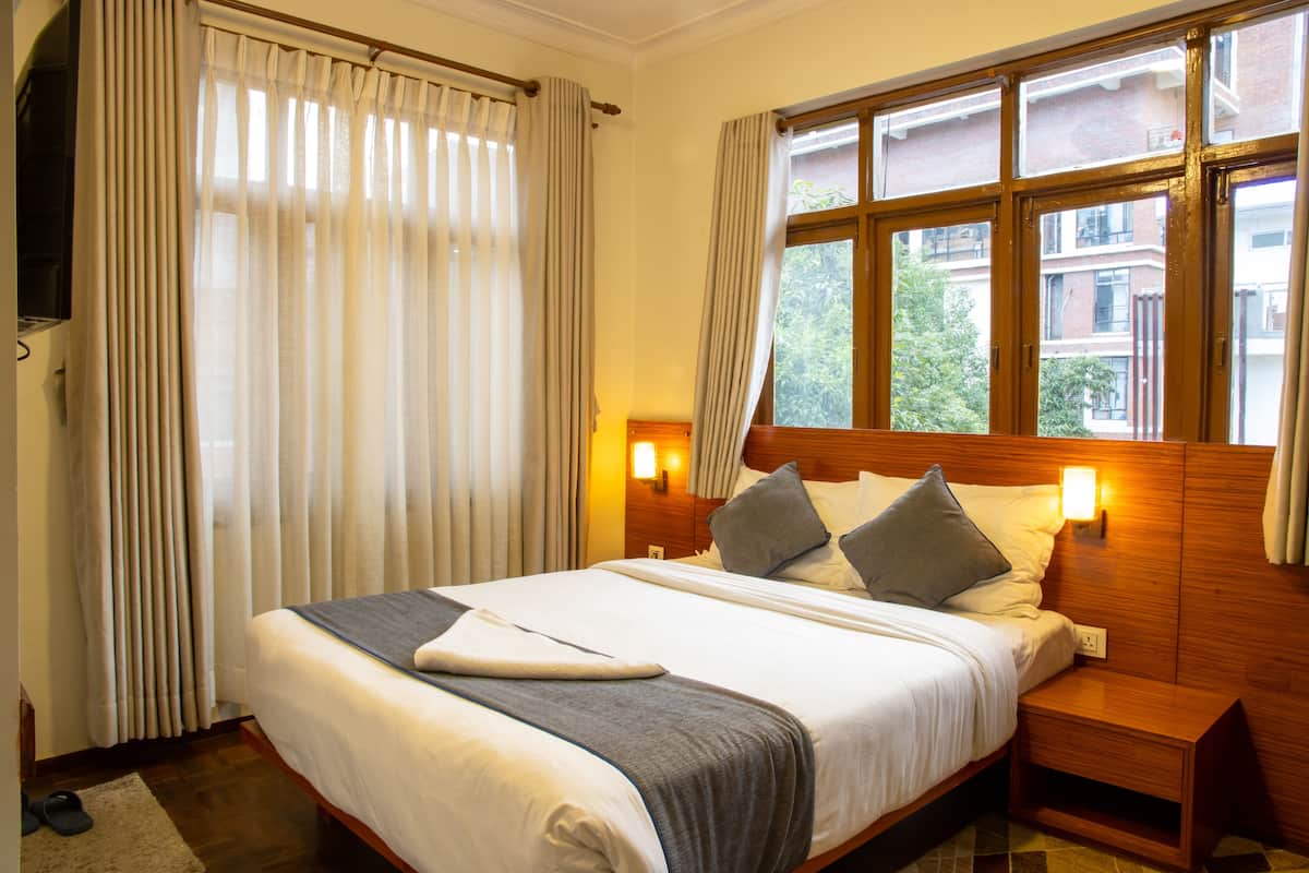 A hotel room with a large bed, wooden headboard, two bedside tables, a window with curtains, and a wall-mounted TV.
