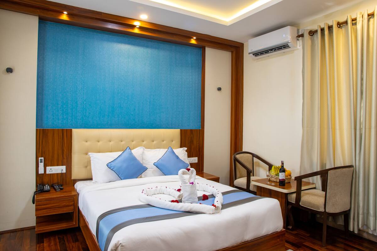 A hotel room with a large bed, a wooden headboard, a bedside table with a phone, and a small table with a bottle and fruit.