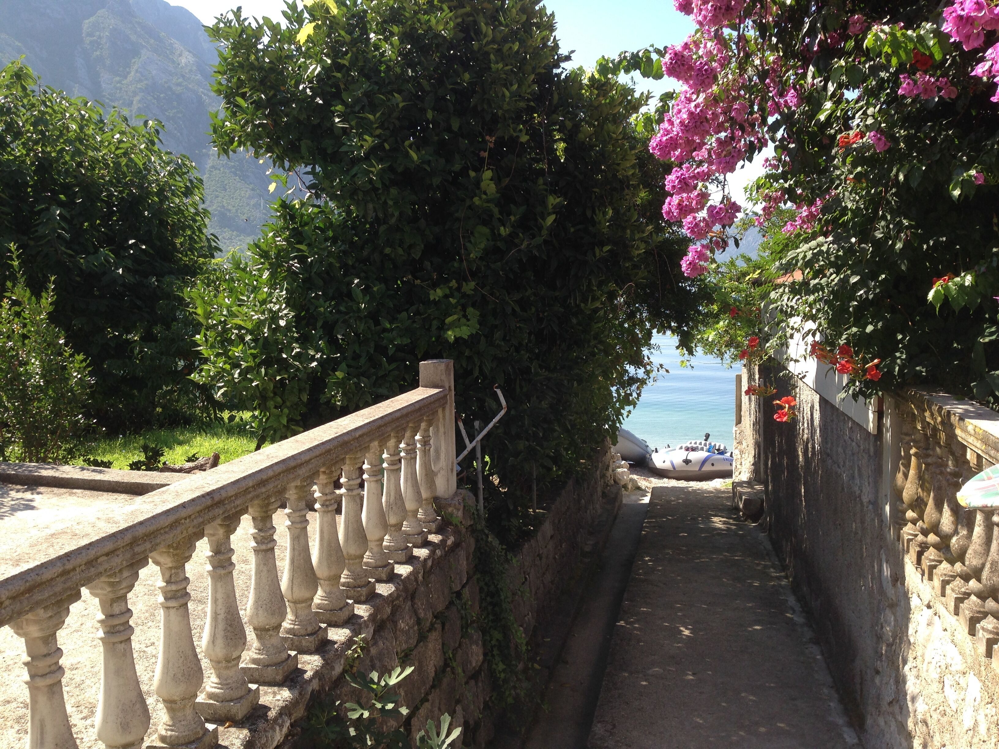 A stone pathway with balustrades, leading to a body of water, surrounded by greenery and mountains.