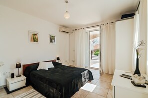 A bedroom with a bed, bedside tables, a desk, a chair, a window with curtains, and an air conditioning unit.