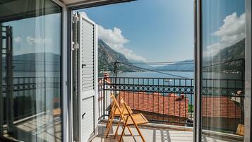 Un balcon avec vue sur la montagne et la mer, une chaise en bois et une rambarde en métal.