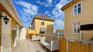 Eine Holzterrasse mit Sitzgelegenheiten im Freien, ein gelbes Gebäude und ein klarer, blauer Himmel.