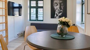 Ein Essbereich mit einem runden Tisch, Stühlen, einer Blumenvase, einem an der Wand montierten Fernseher und einem Fenster mit Blick auf Gebäude.