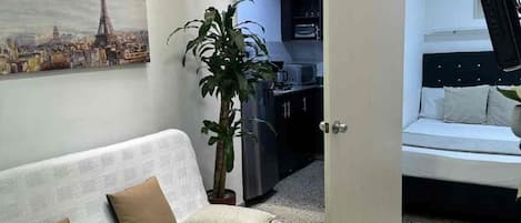 Una sala de estar con sofá, televisión, planta y puerta que da a otra habitación.