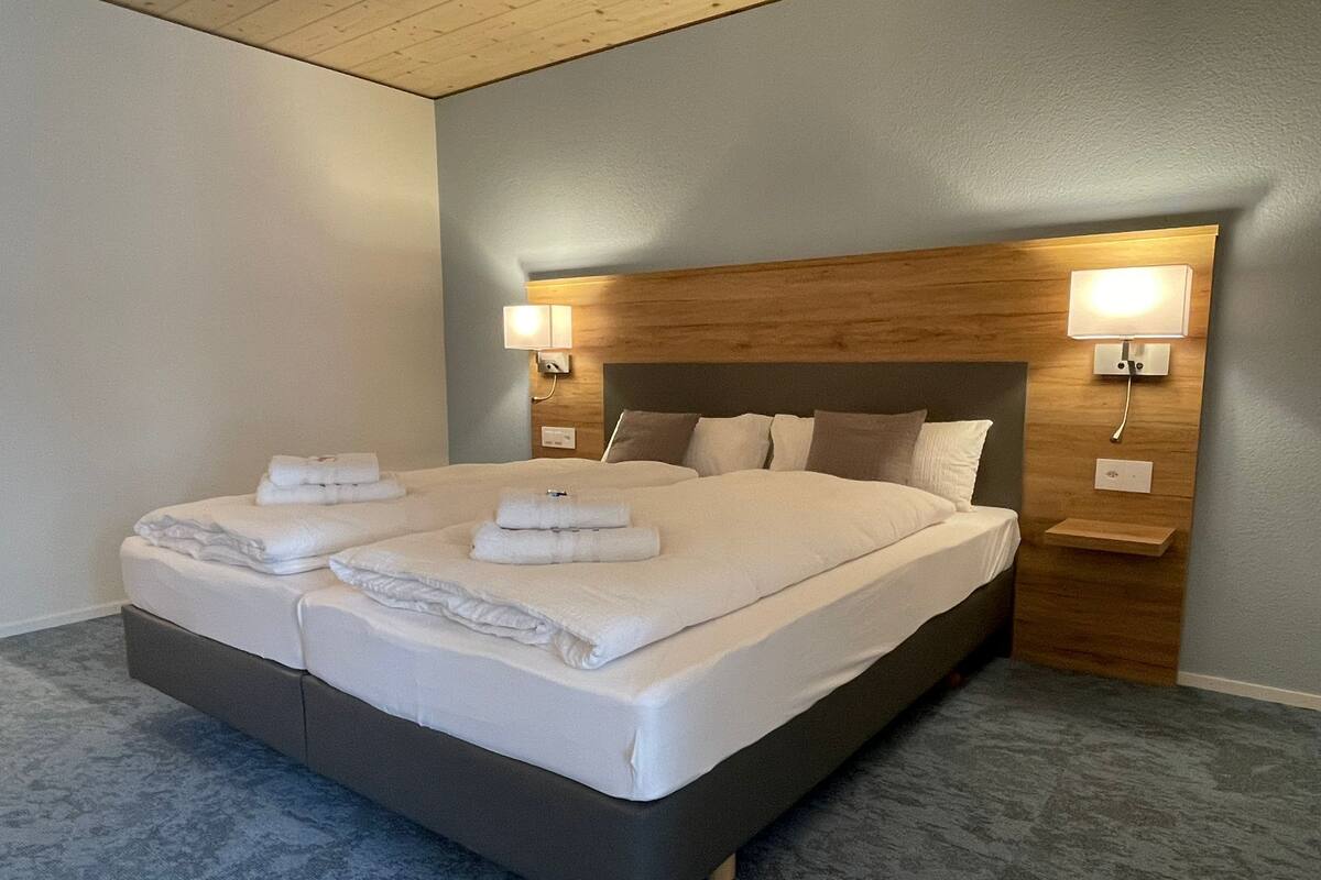 A double bed with white linens and two towels, a wooden headboard, bedside lamps, and a grey carpeted floor.