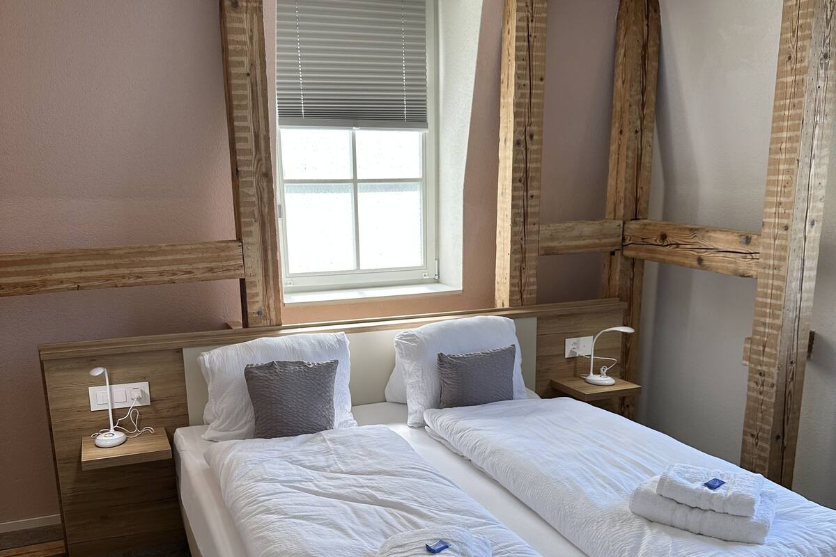 A double bed with white linens and two pillows, a bedside table with a lamp, and a window with blinds.