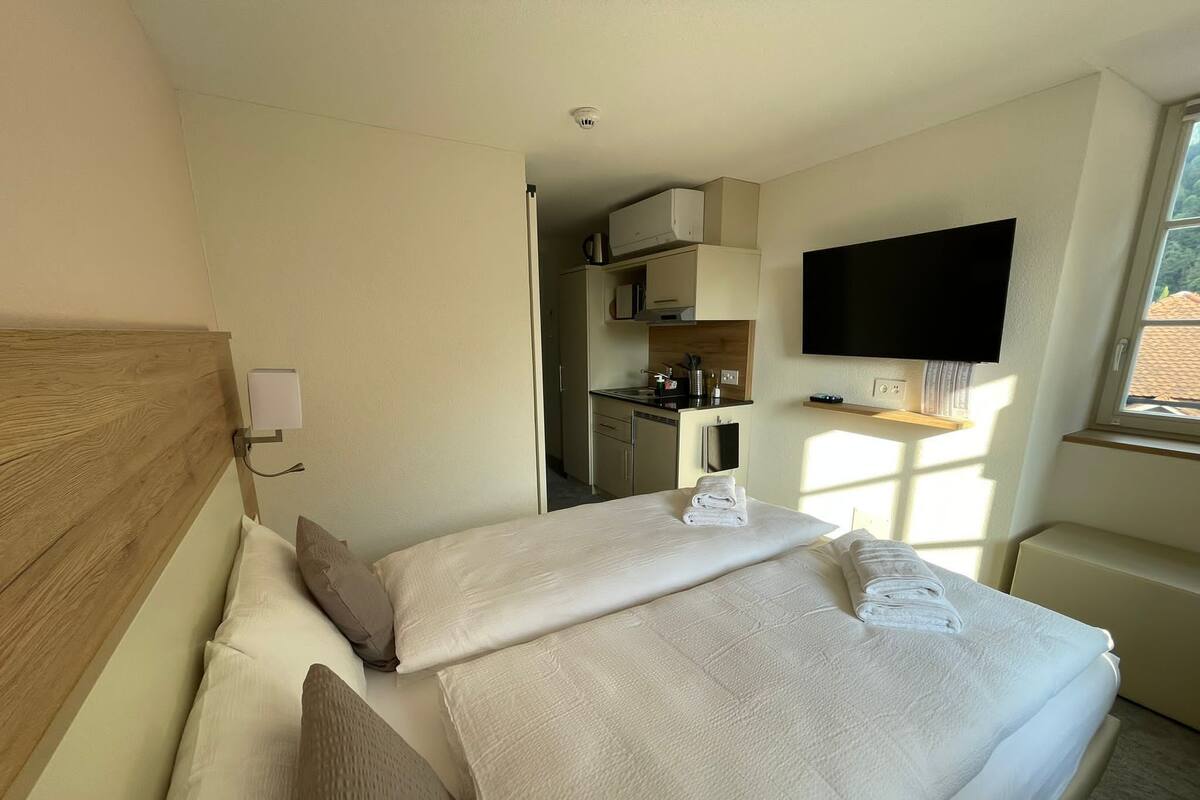A hotel room with a bed, a TV mounted on the wall, and a kitchenette in the background.