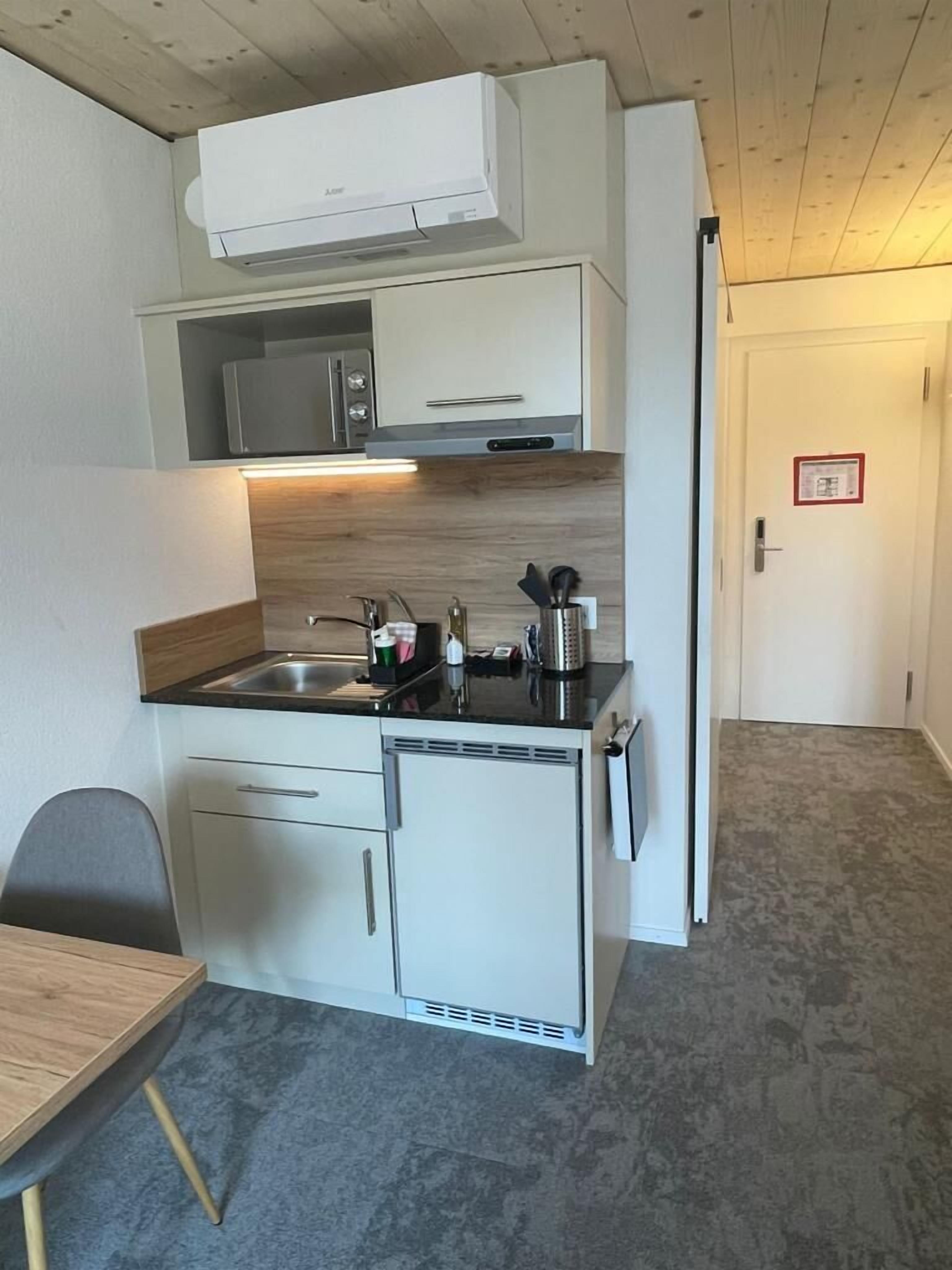 Una kitchenette compacta con microondas, estufa y lavabo, un pequeño comedor con mesa y silla, y una puerta con señal de seguridad contra incendios.