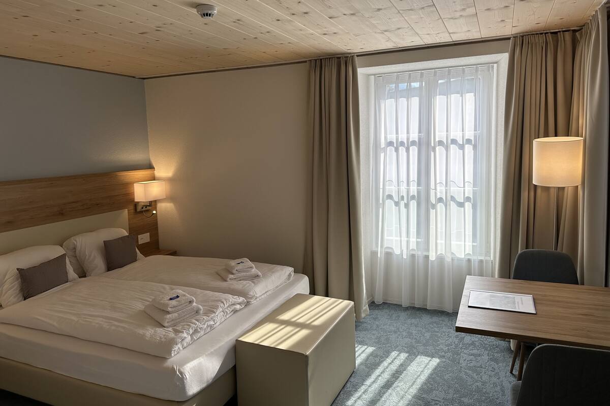 A hotel room with a wooden ceiling, a bed with white linens, a bedside table with a lamp, a desk with a chair, and a window with curtains.