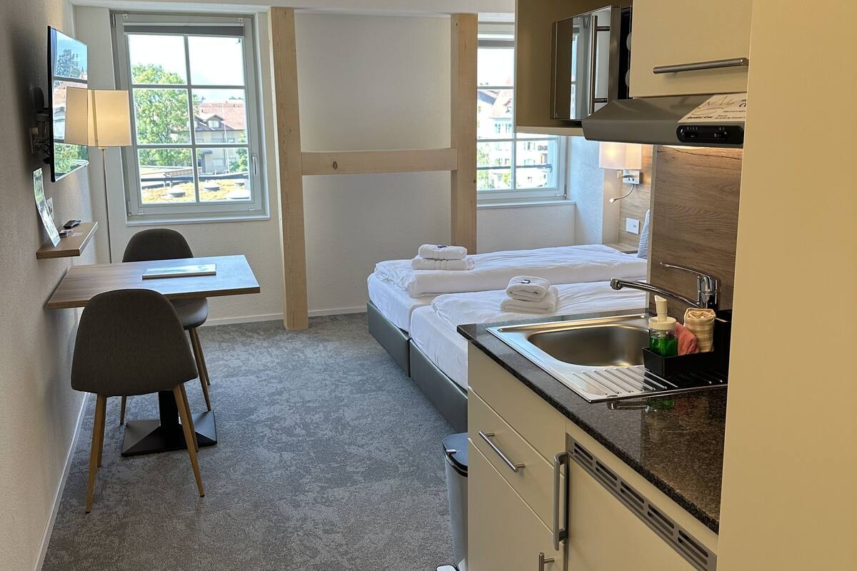 A compact hotel room with a kitchenette, a bed with neatly folded towels, a desk with a chair, and a window with a view of buildings.