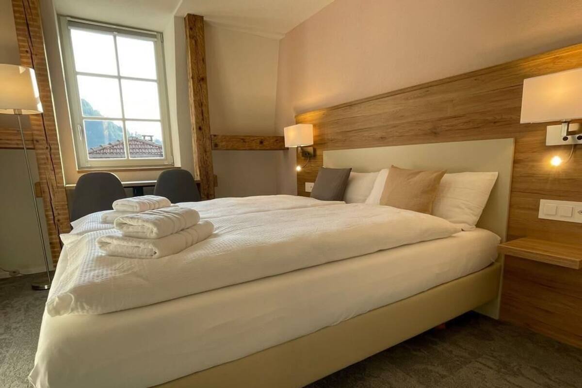 A hotel room with a large bed, two bedside lamps, a desk with a chair, and a window with a view of mountains.