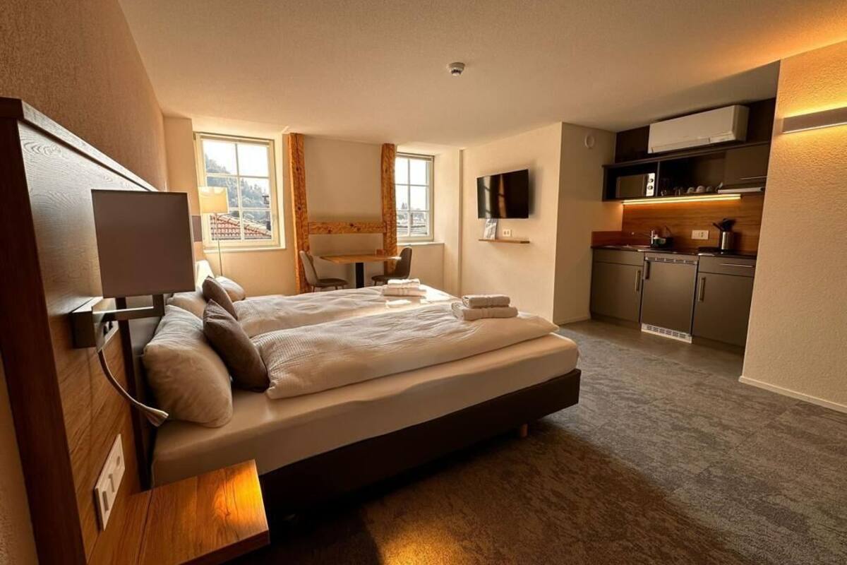 A modern hotel room with a large bed, a TV, a desk, and a kitchenette.