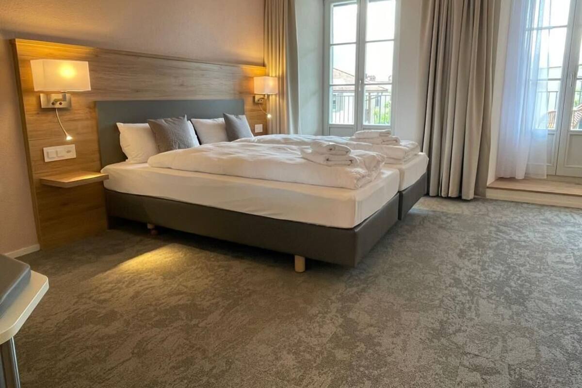 A hotel room with a large bed, two bedside lamps, a wooden headboard, and a view of the outdoors through a window.