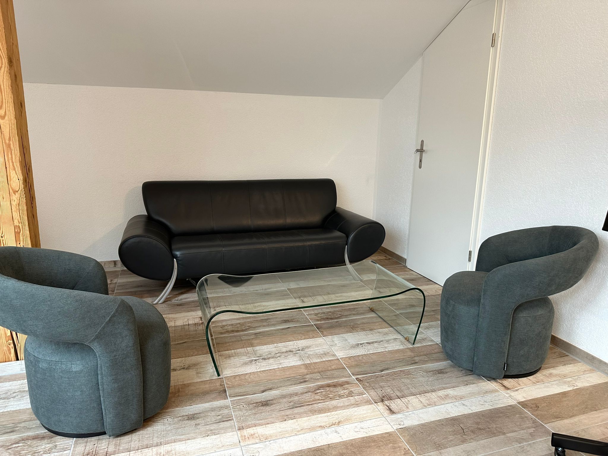 A modern living room with a black leather sofa, two grey armchairs, and a glass coffee table.