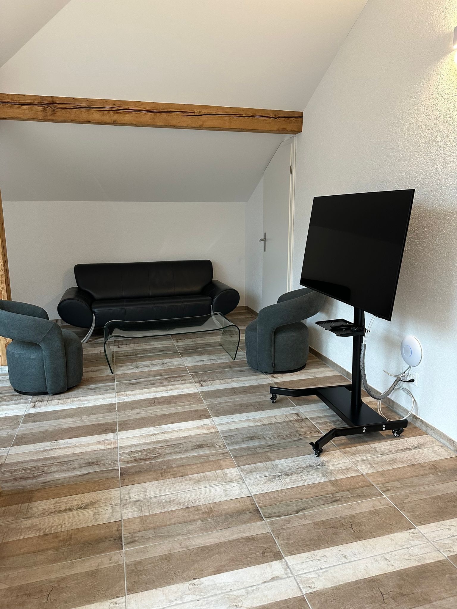 A modern living room with a black sofa, two grey armchairs, a flat-screen TV on a stand, and a wooden ceiling beam.