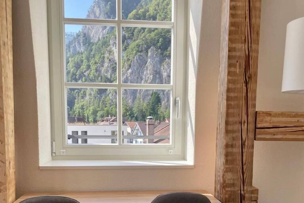 A room with a window overlooking a mountainous landscape, a desk, two chairs, and a lamp.