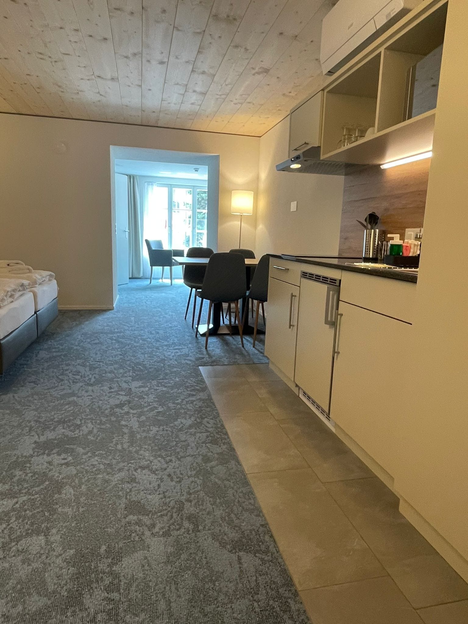 A compact hotel room with a kitchenette, a dining area with chairs, and a large window.