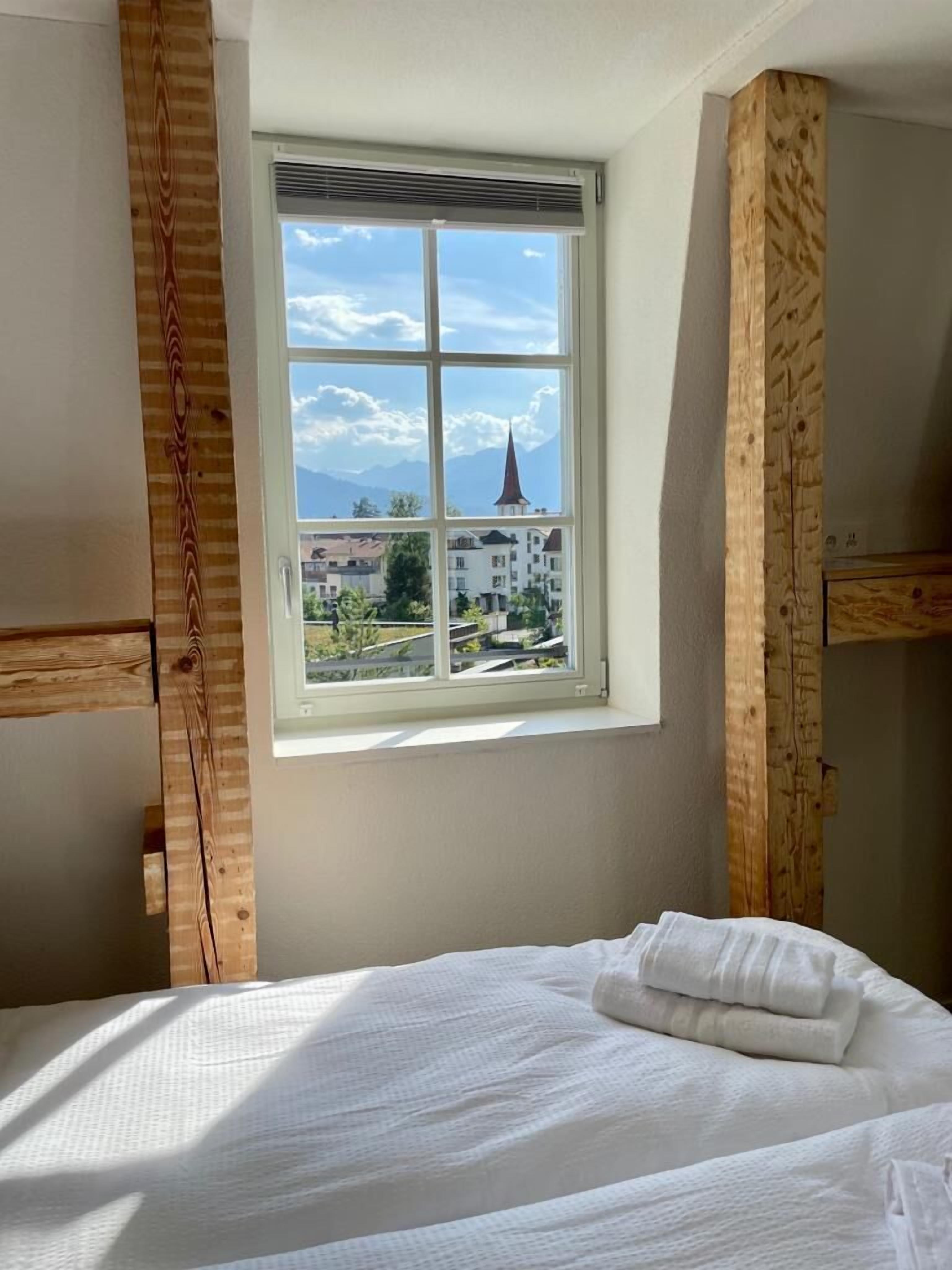 A bedroom with a large window, wooden beams, and a neatly made bed.