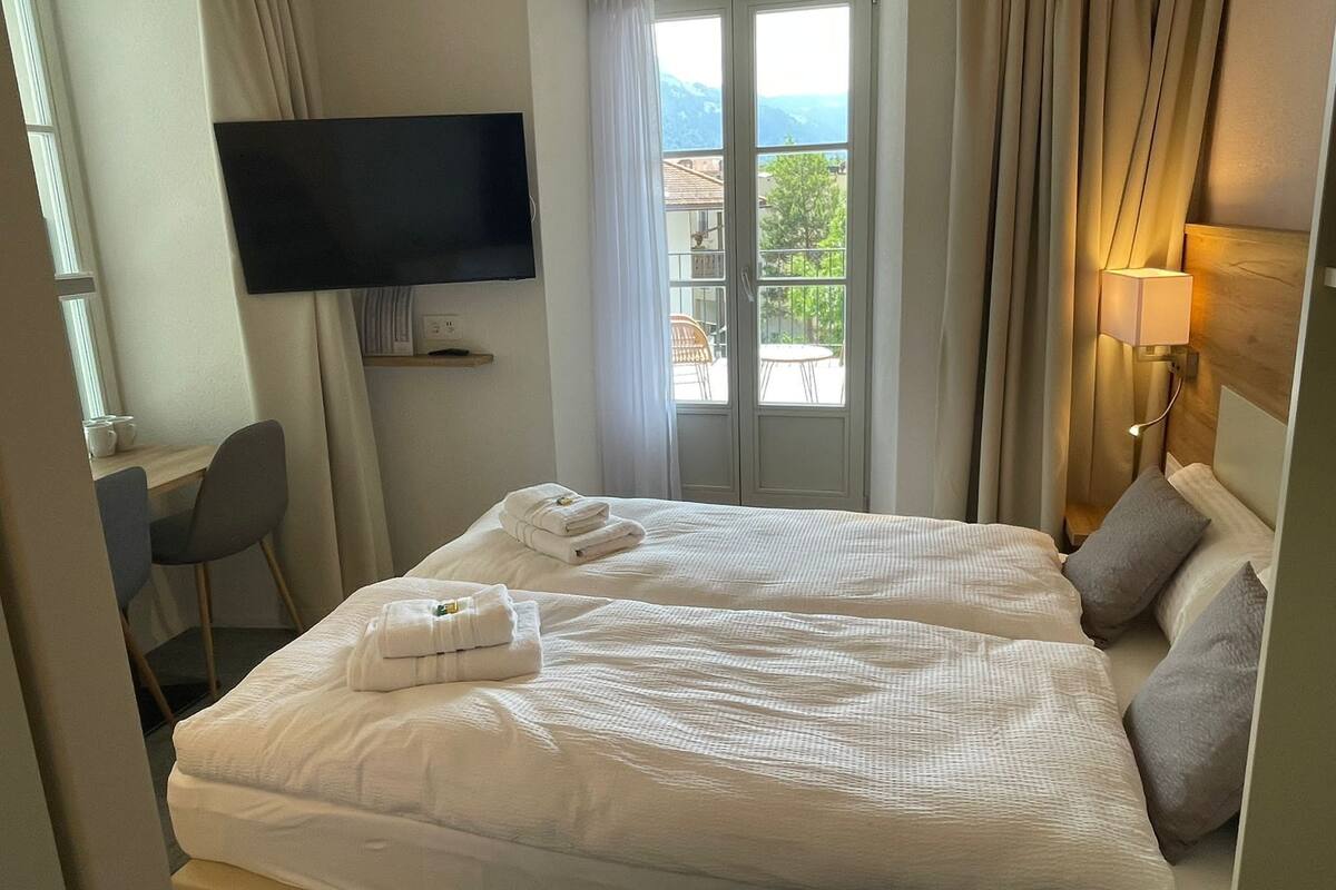 A hotel room with two beds, a TV mounted on the wall, a desk with a chair, and a window with curtains.