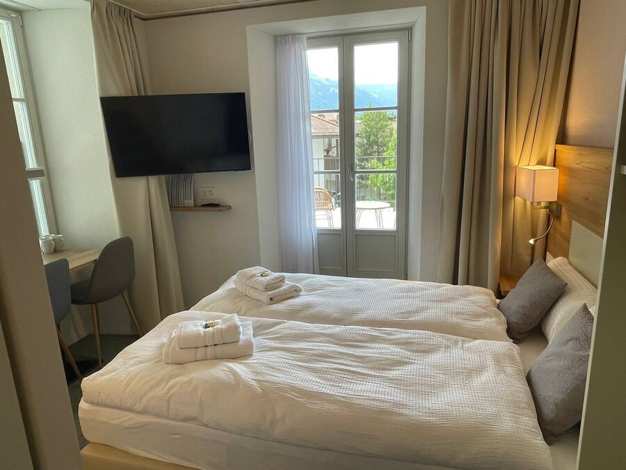 Ein Hotelzimmer mit zwei Betten, einem an der Wand montierten Fernseher, einem Schreibtisch mit Stuhl und einem Fenster mit Vorhängen.