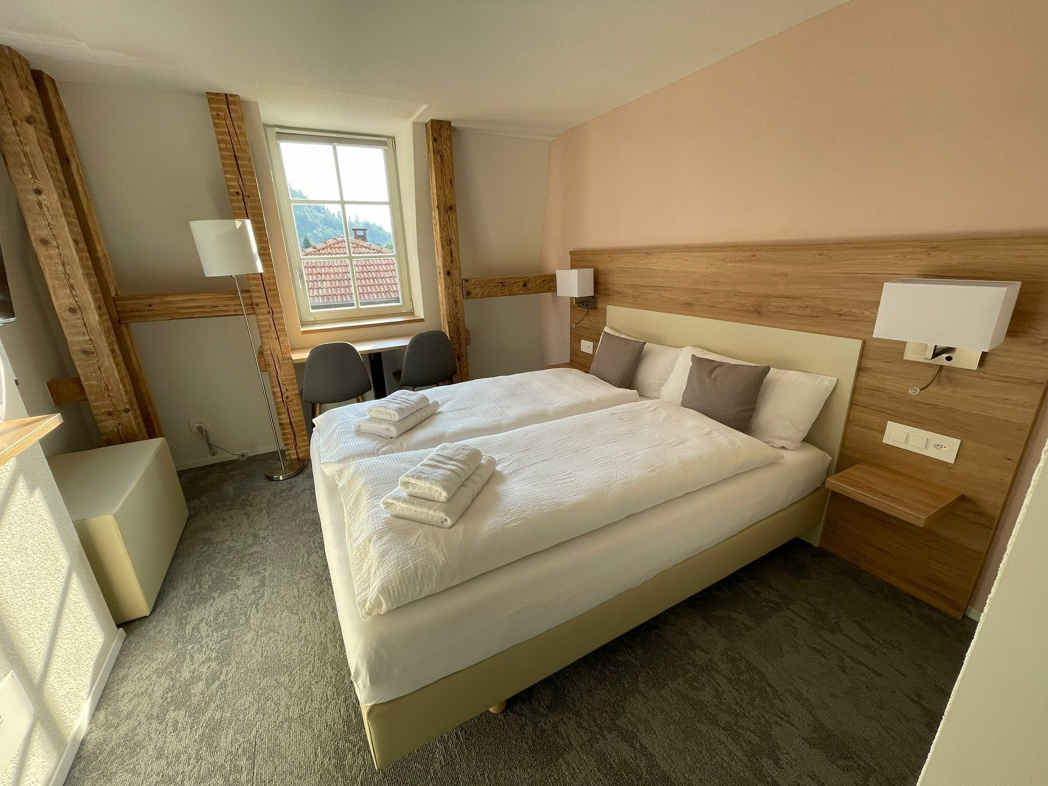 A hotel room with a large bed, two bedside tables, a desk, and a chair. There is a window with a view of buildings.